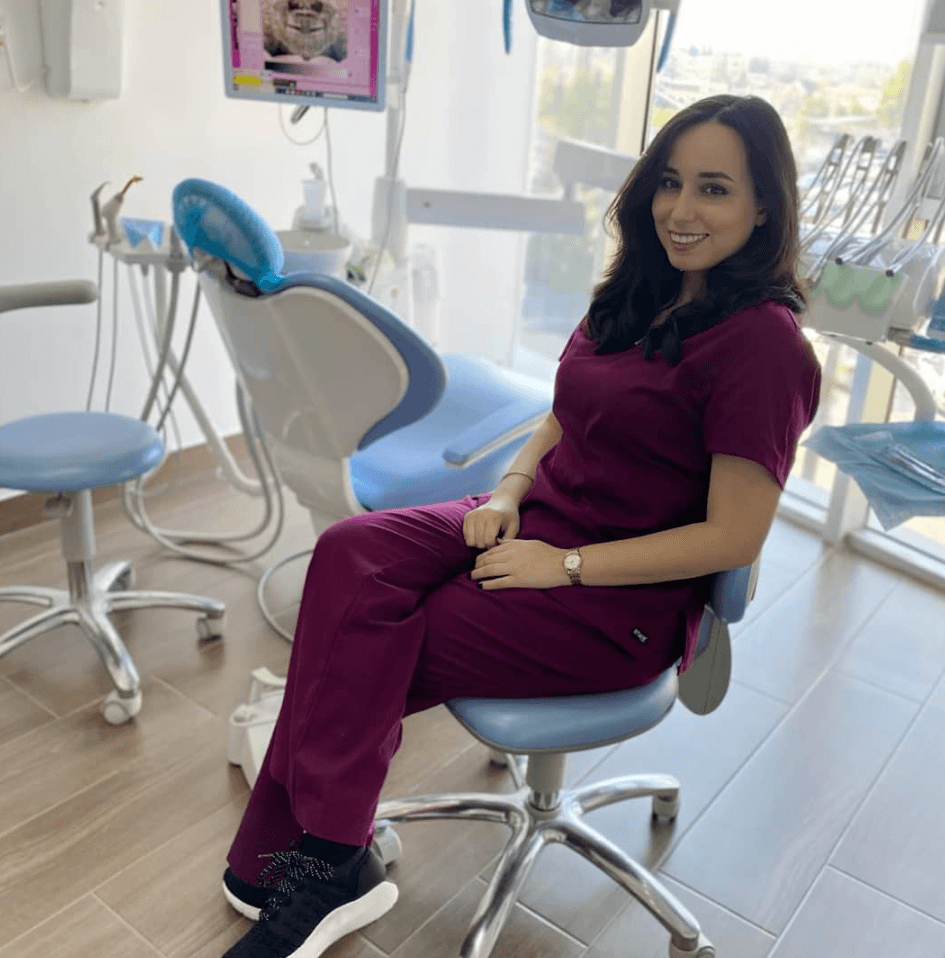 Smiling woman in maroon scrubs sits in a modern dental office with professional equipment.